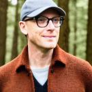 A man wearing glasses, a baseball cap, and a brown sweater standing in a forest.